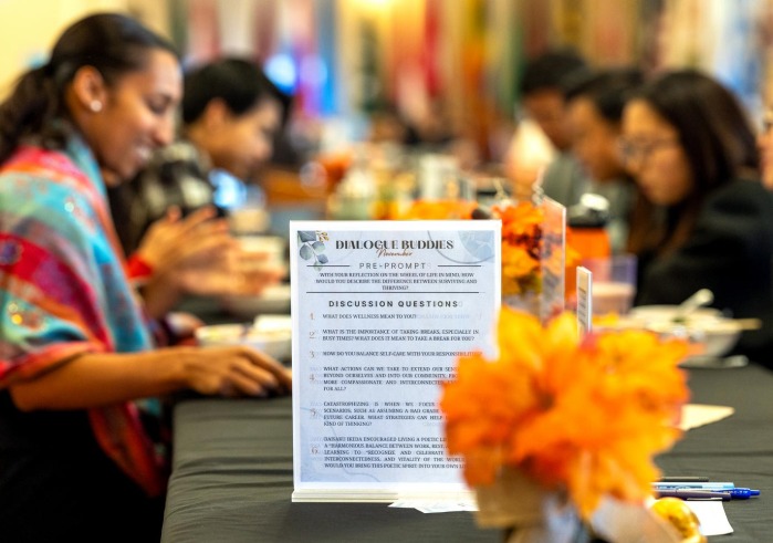 A "Dialogue Buddies" event features reflective questions on wellness and self-care, with participants gathered around a table decorated with orange flowers.