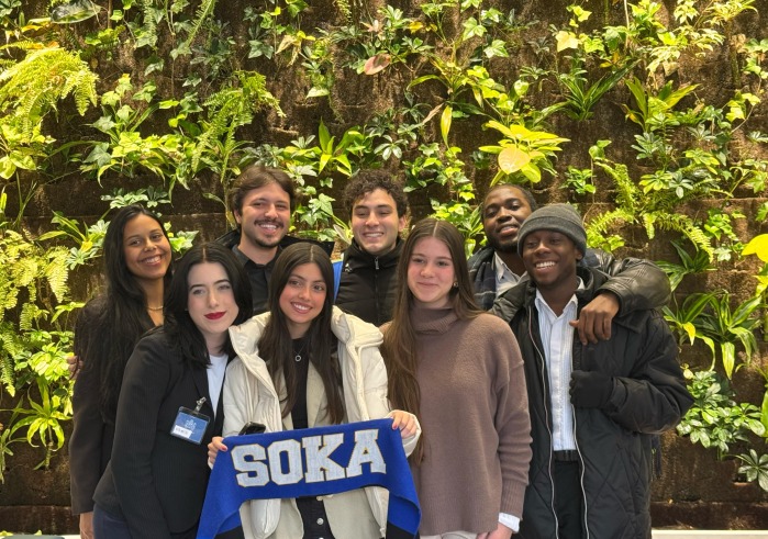 8 student members of the SUA Model U.N. team pose for a group photo in front of a large green wall planter at Toronto's Model U.N. conference. One student is holding up a blue banner that says "SOKA."