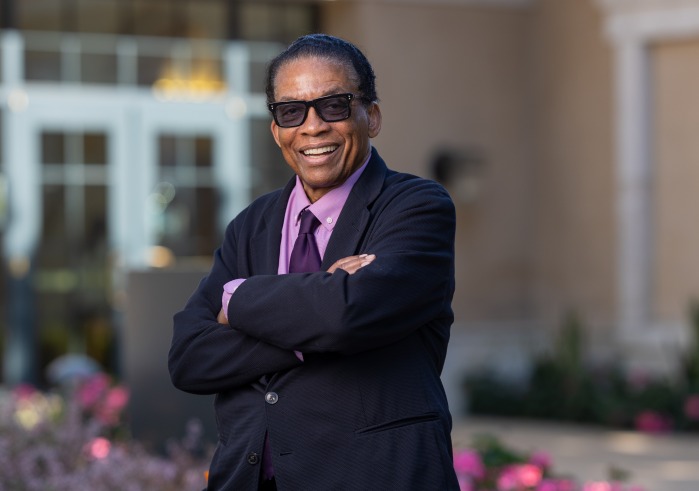 Herbie Hancock wears a black suit jacket over a light purple shirt and a dark purple tie. He is wearing tinted sunglasses and smiles as he stands with his arms crossed outside on Soka's campus.