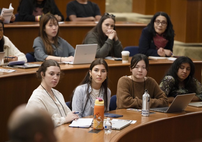 Four students sit alongside eachother at a wooden desk and listen intently to a speaker during the Nuclear Politics 2025 summer program.