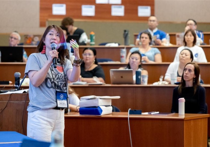 A woman speaks into the microphone and gestures at a projector screen in front of an auditorium of people.