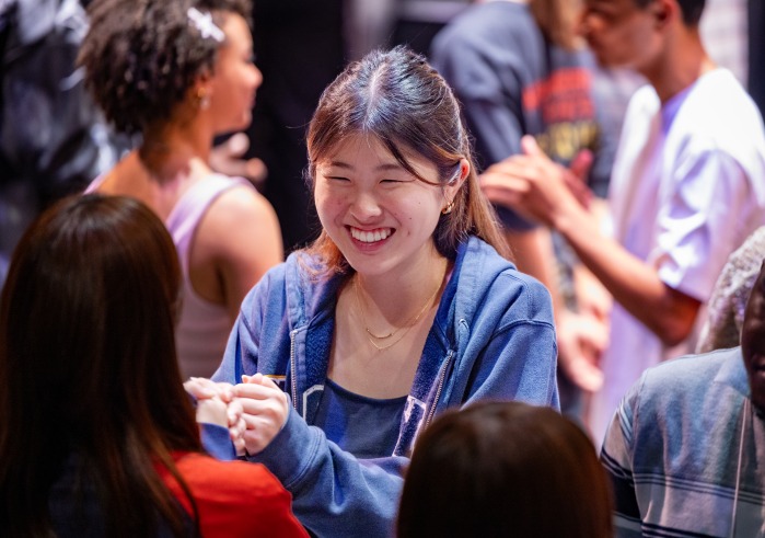 A girl wearing a blue zip-up sweater smiles at another student as they participate in an icebreaker activity.