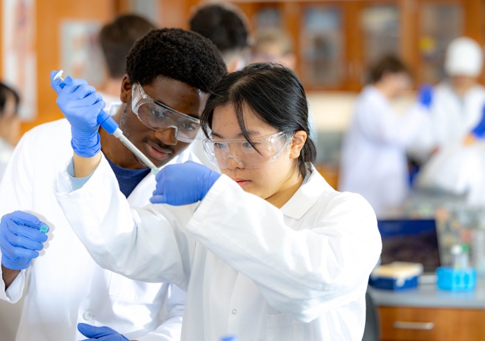 A male and female student in lab coats and safety goggles work together with a micropipette in a science lab.