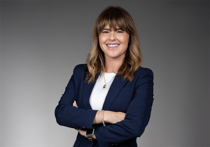 A woman is wearing a navy blazer over a white shirt, with a gold whistle necklace. She is smiling at the camera with her arms crossed.