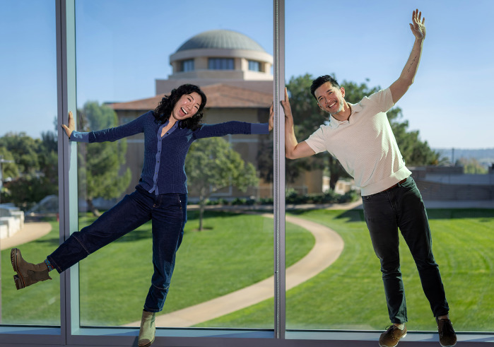 A smiling woman in a blue shirt, jeans, and green boots kicks her right foot and outstretches her arms while a smiling man in a white polo and jeans leans toward her with one arm raised, standing in front of glass windows with Founders Hall behind them.