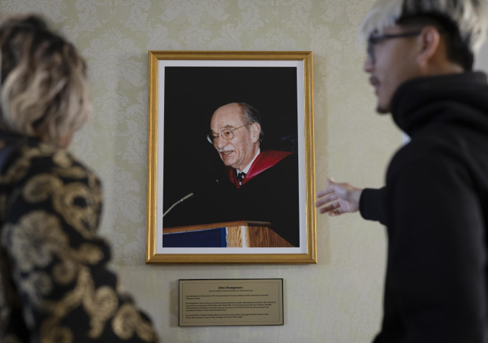 A male student with bleach blonde and black hair gestures towards a framed photo of a man speaking at a lectern.