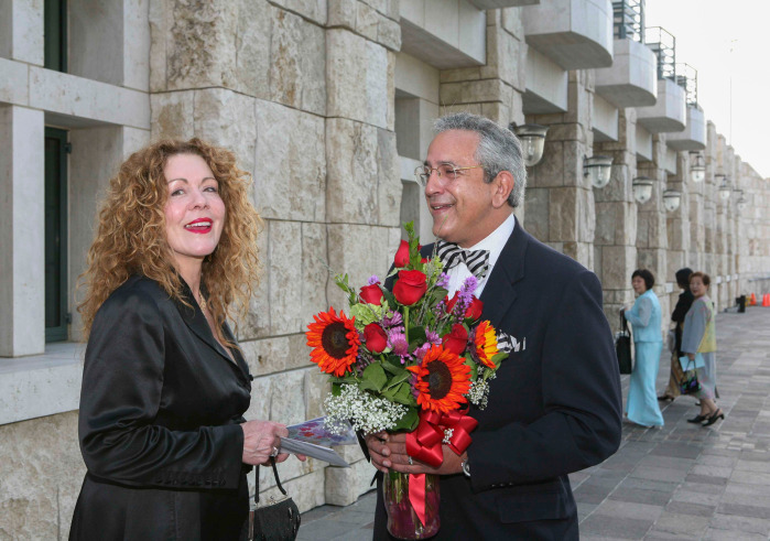Linda and Luis Neieves are wearing black formal attire, smiling, and Luis is holding a bouquet of red and purple flowers as they stand outside of Founders Hall.