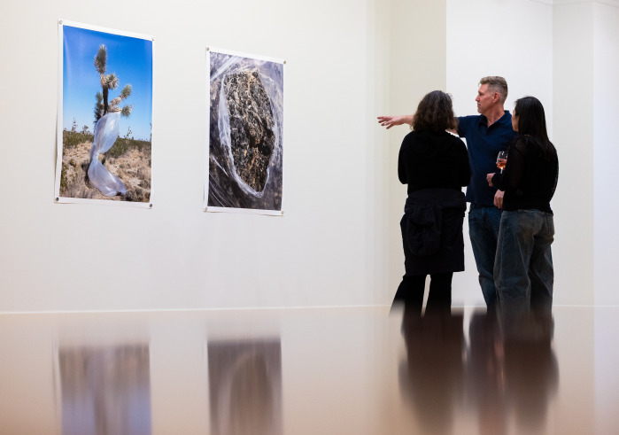 A man wearing a navy polo shirt is gesturing towards two large photographs on a white wall as he explains them to two women wearing black shirts.