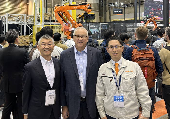 Two men wearing suit jackets and dress shirts and a third man wearing a jacket with "Mujin" embroidered on it smile and pose for a photo together at the 2025 International Robot Exhibition in Tokyo.