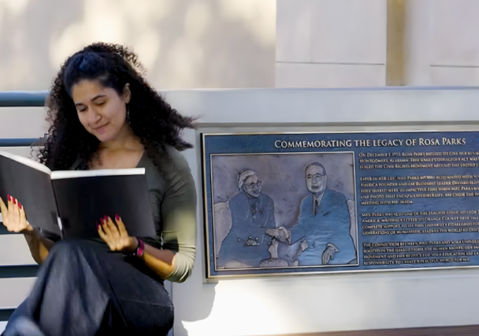 A woman with curly brown hair looks at a large black book as she sits beside a plaque commemorating Rosa Parks' legacy.