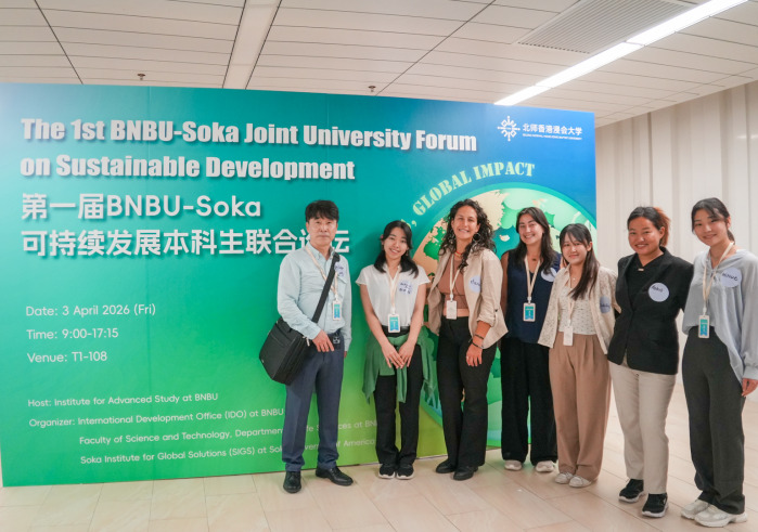A man stands next to six women, all wearing nametags, as they pose in front of a large green and blue sign reading "The 1st BNBU-Soka Joint University Forum on Sustainable Development"