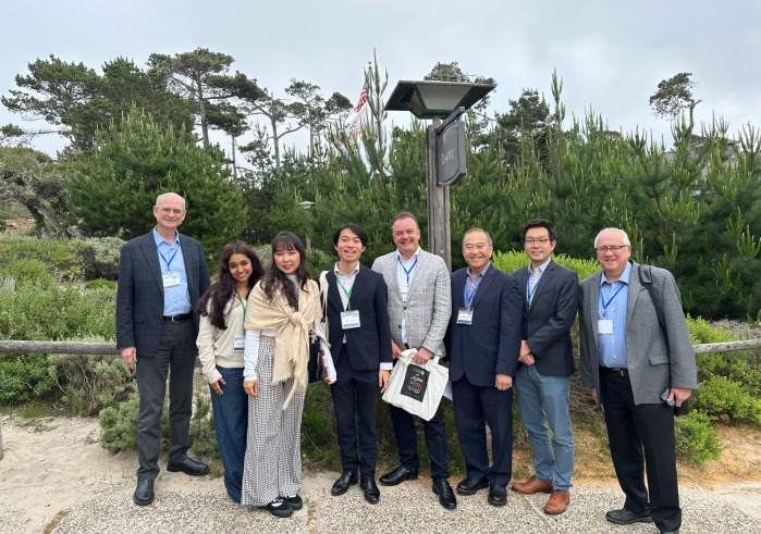 A group of conference attendees wearing formal attire pose outside in front of various greenery on Asilomar Conference Grounds in Pacific Grove, California.