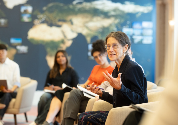 A woman with brown hair in a ponytail and wearing glasses speaks with her hands. Out-of-focus students are in the background listening sitting in front of a wall-sized navy, gold, and green world map.