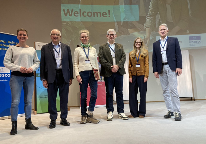 Six conference attendees and presenters stand shoulder to shoulder in front of a projector screen that says "Welcome!"