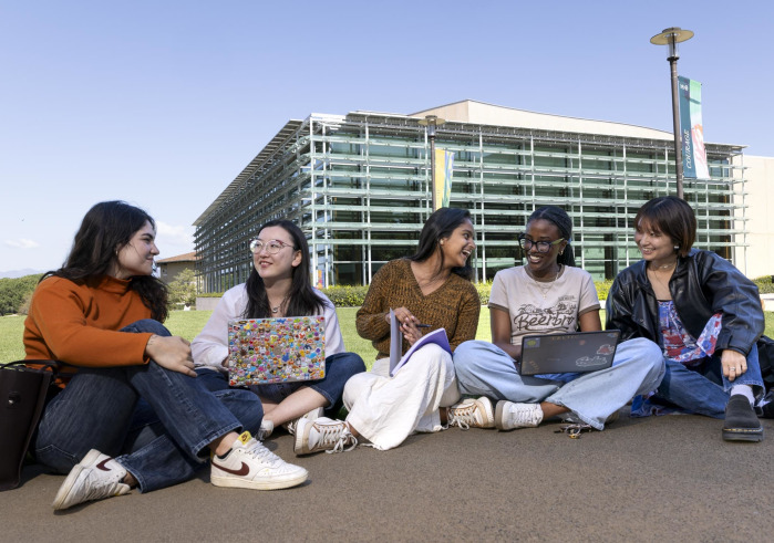 Five female students sit on the sidewalk in front of the Performing Arts Center, some with laptops in their laps, as they smile and laugh with eachother.