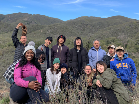 Twelve students and their professor pose together in front of green rolling hills.