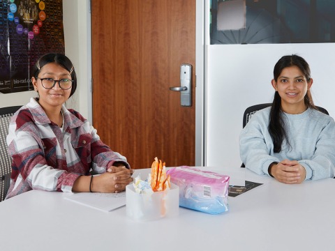 Two women smile at the camera as they sit at a table with menstrual products