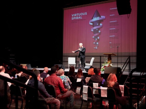 Simon Mainwaring holds a microphone and stands on stage in the Black Box Theatre on Soka's campus where he speaks to an audience about Rewiring Capitalism. There is a screen behind projecting some of his presentation points.