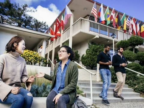 Two female students talk while sitting on a bench beneath a building displaying flags from various countries. Two men are walking down the steps to the side of the women talking.