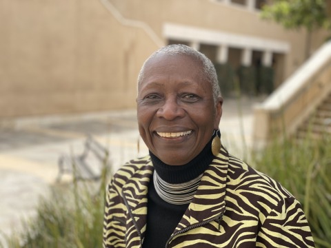 Professor Emeritus Gail Thomas smiles outside on Soka University's campus. She's wearing a black turtleneck with a zebra print zip up jacket and gold dangly earrings.