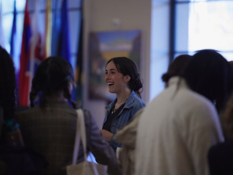 A brunette woman wearing a jean jacket smiles as she speaks with other students in front of a variety of international flags.