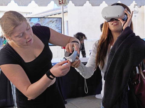 A woman wearing a VR headset is assisted by another woman.