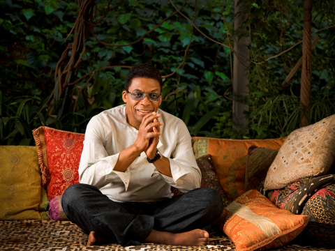 Herbie Hancock sits crosslegged surrounded by colorful throw pillows. He is clasping his hands and smiling at the camera while wearing a white striped shirt, dark jeans, and tinted glasses. Photo by Douglas Kirkland