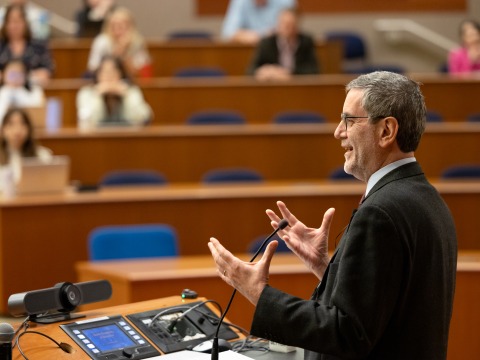 Andrea Bartoli, wearing a suit and glasses, stands at a lectern and gestures with his hands while speaking to an auditorium of people.
