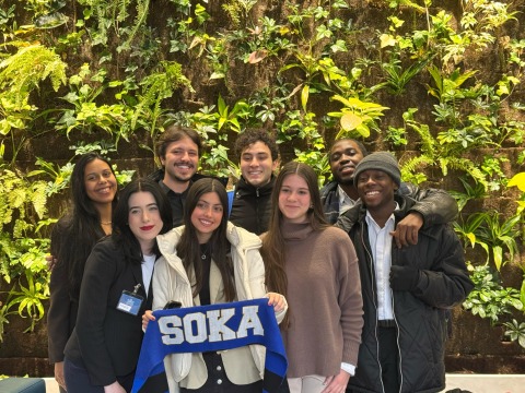 8 student members of the SUA Model U.N. team pose for a group photo in front of a large green wall planter at Toronto's Model U.N. conference. One student is holding up a blue banner that says "SOKA."