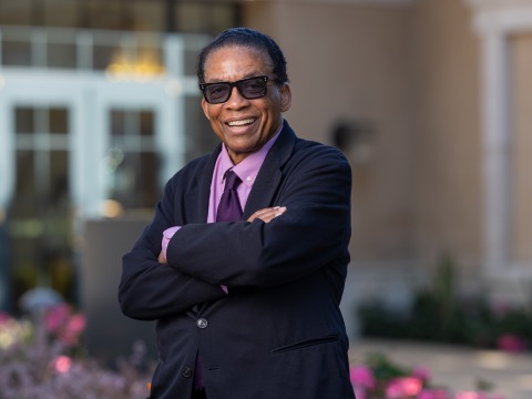 Herbie Hancock wears a black suit jacket over a light purple shirt and a dark purple tie. He is wearing tinted sunglasses and smiles as he stands with his arms crossed outside on Soka's campus.