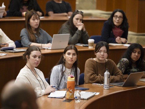Four students sit alongside eachother at a wooden desk and listen intently to a speaker during the Nuclear Politics 2025 summer program.