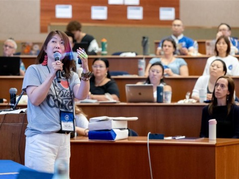 A woman speaks into the microphone and gestures at a projector screen in front of an auditorium of people.