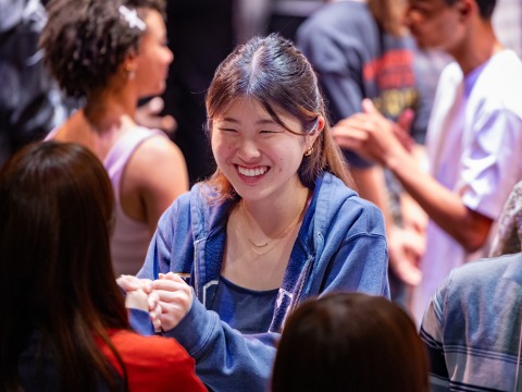 A girl wearing a blue zip-up sweater smiles at another student as they participate in an icebreaker activity.