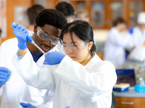 A male and female student in lab coats and safety goggles work together with a micropipette in a science lab.