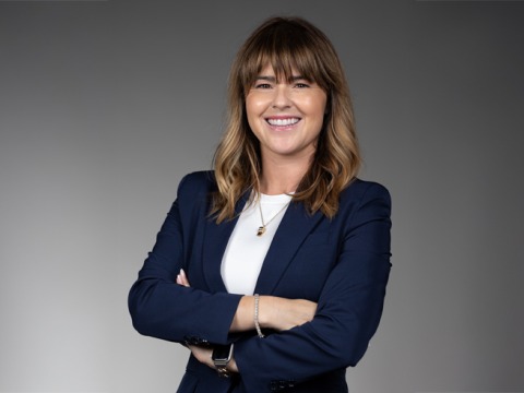 A woman is wearing a navy blazer over a white shirt, with a gold whistle necklace. She is smiling at the camera with her arms crossed.