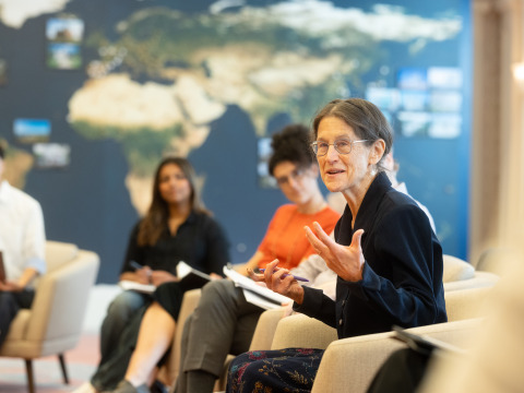 A woman with brown hair in a ponytail and wearing glasses speaks with her hands. Out-of-focus students are in the background listening sitting in front of a wall-sized navy, gold, and green world map.