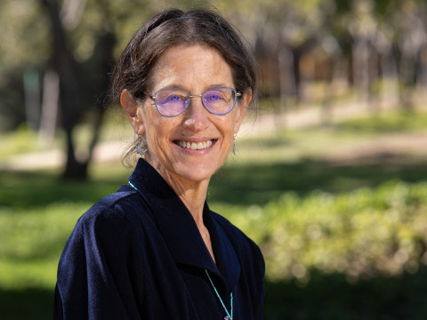 A dark-haired woman wearing a navy blue collared shirt and blue-rimmed glasses smiles at the camera outside in a grassy area.