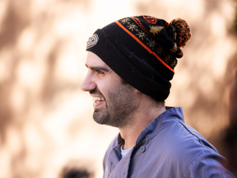 Side profile shot of a bearded man wearing a grey chef shirt and a black Anaheim Ducks holiday beanie smiles.