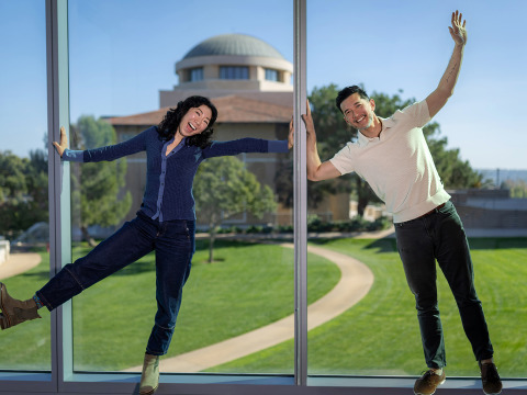 A smiling woman in a blue shirt, jeans, and green boots kicks her right foot and outstretches her arms while a smiling man in a white polo and jeans leans toward her with one arm raised, standing in front of glass windows with Founders Hall behind them.