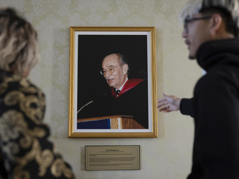 A male student with bleach blonde and black hair gestures towards a framed photo of a man speaking at a lectern.