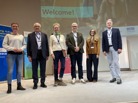 Six conference attendees and presenters stand shoulder to shoulder in front of a projector screen that says "Welcome!"