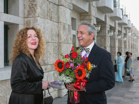 Linda and Luis Neieves are wearing black formal attire, smiling, and Luis is holding a bouquet of red and purple flowers as they stand outside of Founders Hall.