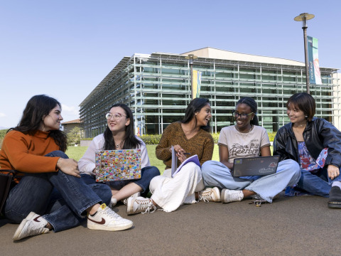 Five female students sit on the sidewalk in front of the Performing Arts Center, some with laptops in their laps, as they smile and laugh with eachother.
