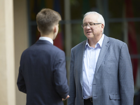 SUA President Ed Feasel is wearing a grey suit jacket and white shirt. He smiles as he speaks to a student wearing a blue suit. 