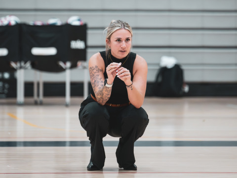 A blonde woman with her hair up, wearing a black tanktop and pants squats down on a gym floor with volleyballs in the distance.