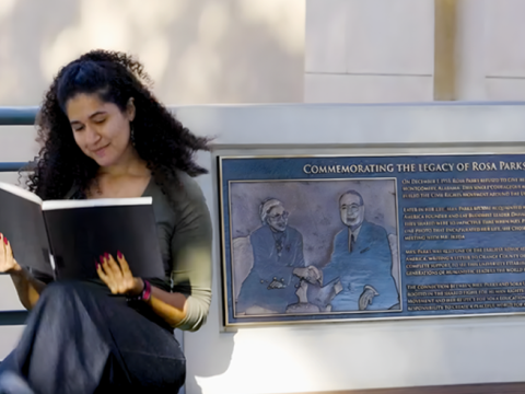 A woman with curly brown hair looks at a large black book as she sits beside a plaque commemorating Rosa Parks' legacy.