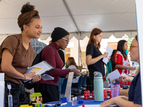 A young woman with a brown headwrap and gold jewelry smiles while leaning over a table at an outdoor career fair, holding a clipboard and documents. Other students and recruiters are visible under a large white tent in the background.
