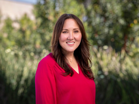 A professional headshot of a smiling woman with long brown hair, wearing a vibrant magenta V-neck blouse. She is positioned against a soft-focus background of lush green foliage and trees.