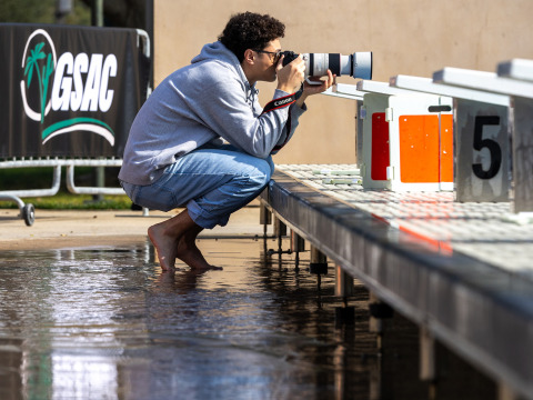 A barefoot photographer in a grey hoodie and jeans crouches on a wet pool deck, aiming a Canon camera with a large telephoto lens toward the starting blocks. In the background, a banner reads "GSAC."
