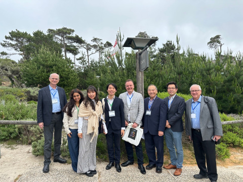 A group of conference attendees wearing formal attire pose outside in front of various greenery on Asilomar Conference Grounds in Pacific Grove, California.